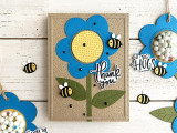 Handmade greeting card with flower on it and thank you sentiment on it with bumble bees and crafting materials in background.