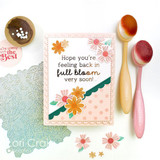 Handmade greeting card with flowers and dots on it and hope you're feeling back in full bloom very soon sentiment on it with crafting materials in background.