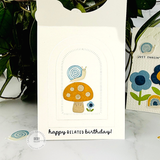 Handmade greeting card with a mushroom, snail, and flower on it with happy belated birthday sentiment on it with cards in background.