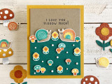 Handmade greeting card with mushrooms, flowers, and snails with I love you slow much sentiment on it with mushrooms, flowers, and snails in background.