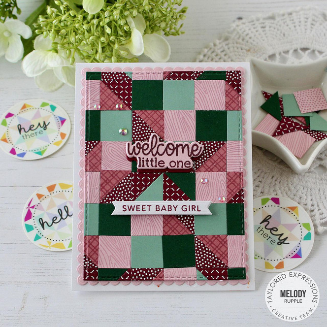 Handmade greeting card with patterned squares on it and welcome little on and sweet baby girl sentiments on it with crafting materials in background.