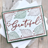 Handmade greeting card featuring a foiled Grateful sentiment layered over a soft botanical background with green and orange accents.
