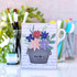 Handmade greeting card with flowers in a pail with hello sentiment on it and crafting materials in background.