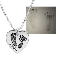 Close up of actual footprints on a silver heart necklace on a white background, shown with the original footprints. The baby's name is stamped on the front of the charm