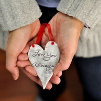 Model holding personalized fine pewter heart Christmas ornament, engraved with husband's handwriting, hung on a red ribbon