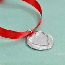 Front side of the Love You Handwritten Christmas Tree Ornament, featuring a hand sculpted fine pewter disc with a raised outline of a heart, hung on a red ribbon. Shown from the side on green background