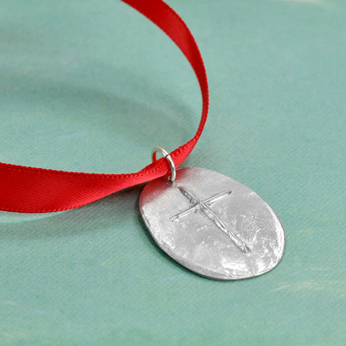 Front of custom fine pewter Cross Ornament, featuring a raised cross on a hand sculpted oval, hung on a red ribbon, shown from the side on a green background.