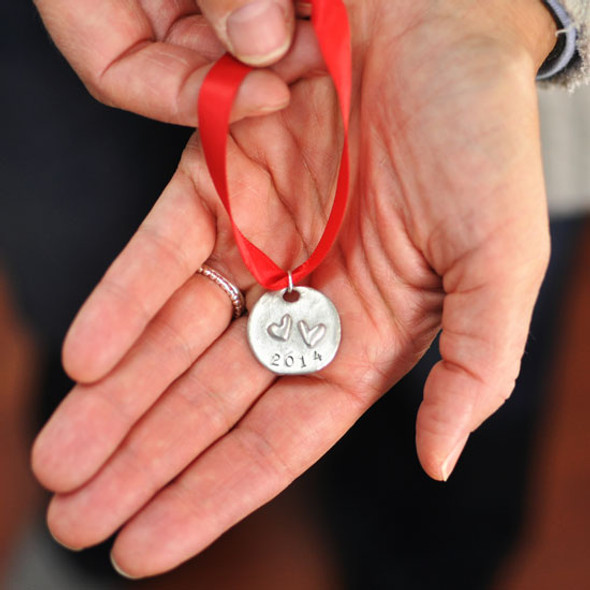 Model holding hand stamped double heart ornament. The ornament is made with fine pewter, hung on a red ribbon, with 2 raised hearts, and the year stamped on the bottom.