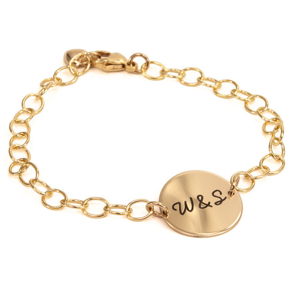 Personalized gold disc bracelet, with the disc custom engraved with husband's handwritten "W&S" for an anniversary present. Charm is on a gold chain, with a gold puffed heart charm attached to the clasp. Shown from the front at a low angle, on a white background