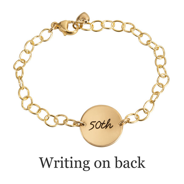 Back of personalized gold disc bracelet, with the disc custom engraved with husband's handwritten "50th" for an anniversary present. Charm is on a gold chain, with a gold puffed heart charm attached to the clasp. Shown from the back on a white background