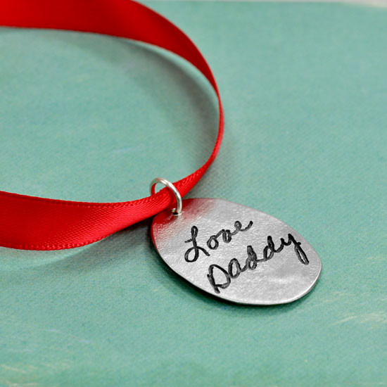 Back of custom handwriting Cross Ornament, personalized with late father's actual handwritten signature engraved on a fine pewter oval, hung on a red ribbon, shown from the side on a green background.