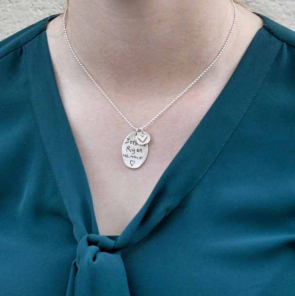 Model wearing handmade sterling silver oval custom engraved with kids' actual signatures. Paired with silver disc with silver raised heart, hung on a silver ball chain.