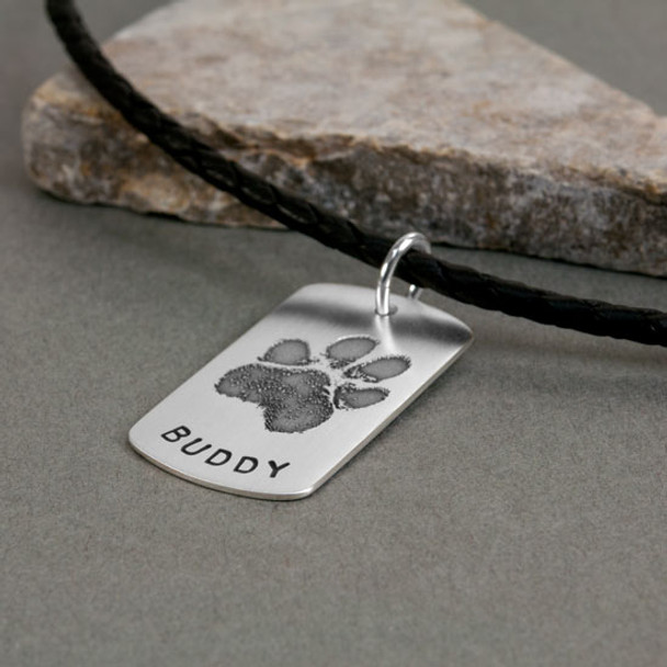 Custom sterling silver military dog tag, personalized with pet's pawprints, and stamped with pet's name "Buddy". Shown from the side on gray paper next to a rock