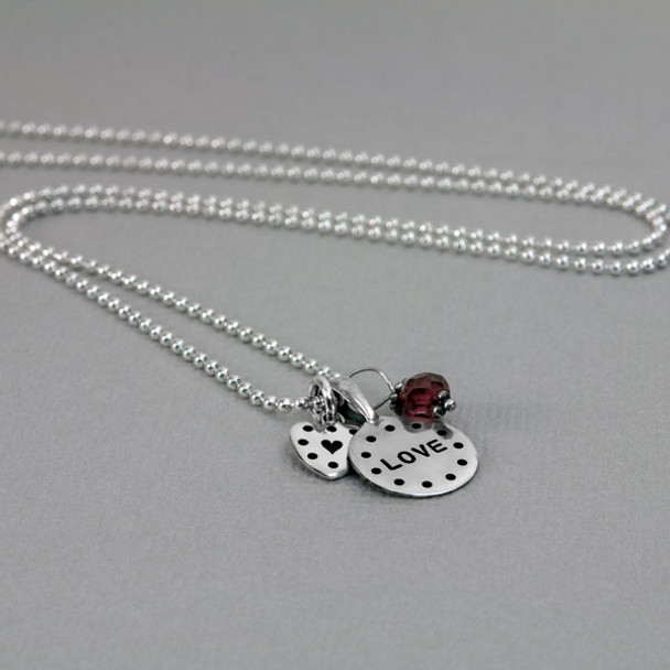 Side view of personalized silver disc and silver heart necklace, hand stamped with dotted border and the word love, with a garnet birthstone, shown close up on white