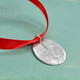 Front of custom fine pewter Cross Ornament, featuring a raised cross on a hand sculpted oval, hung on a red ribbon, shown from the side on a green background.