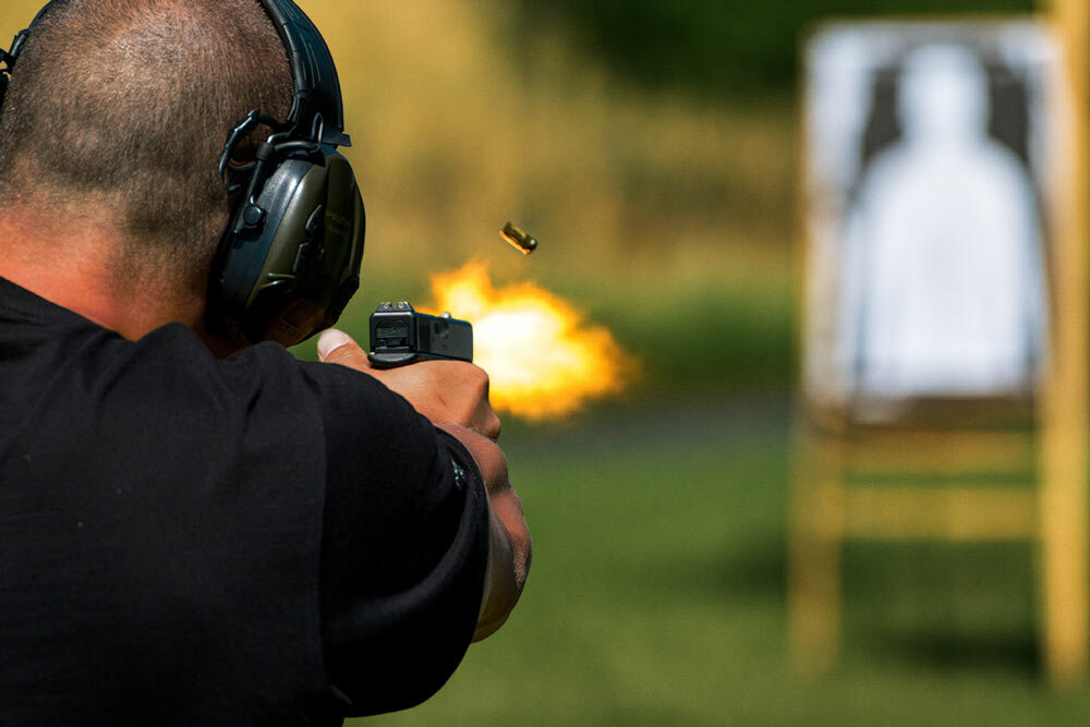 Person wearing hearing protection fires a handgun; muzzle flash and spent casing caught mid-air, with a blurred silhouette target downrange.