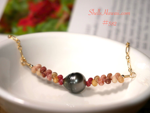 An elegant coastal Hawaiian shell necklace resting in a white dish, highlighting the natural color flow of red-hot pink, yellow Kamoa green, soft pink, brown, and off-white Kahelelani shells with a 9.5 mm Tahitian pearl.