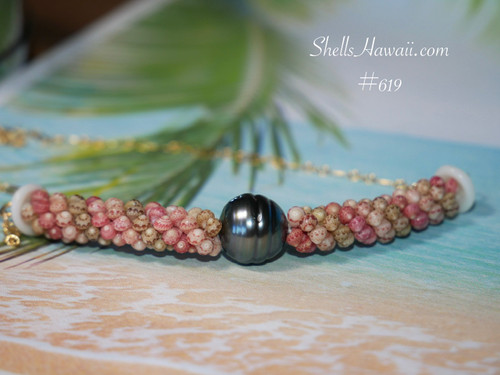 Front lay flat view of the triple strand poepoe necklace displayed on a tropical beach-style background highlighting the balanced arrangement of pink, green, and keʻokeʻo Kahelelani shells with the baroque peacock Tahitian pearl centered in the design.