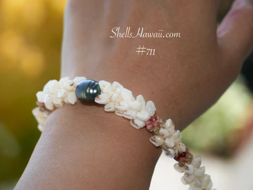 nibl_711
Close-up of a Niʻihau shell bracelet worn on the wrist, featuring a lustrous dark pearl surrounded by off-white Momi shells and colorful Kahelelani shell accents.