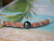 Front lay flat view of the triple strand poepoe necklace displayed on a tropical beach-style background highlighting the balanced arrangement of pink, green, and keʻokeʻo Kahelelani shells with the baroque peacock Tahitian pearl centered in the design.