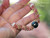Angled close-up handheld view showing the texture, shell placement, and detailed triple strand weaving of pink, green, and keʻokeʻo Kahelelani shells around the baroque peacock Tahitian pearl centerpiece.