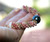 Side angle close-up of the necklace resting across a hand highlighting the thickness, structure, and rounded poepoe weaving pattern created with pink, green, and keʻokeʻo Kahelelani shells and a baroque peacock Tahitian pearl.