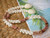 Handmade Kipona Style Hawaiian shell necklace displayed beside a tropical beach postcard, showcasing colorful Kahelelani shells, off-white Momi shells, and a traditional Granulated Cowrie shell clasp.