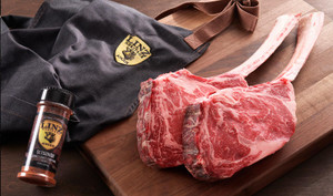 Pictured is 2 uncooked dry aged tomahawk ribeyes on a cutting board along side an apron and Linz Heritage Angus seasoning