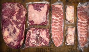 Pictured are uncooked and individually wrapped USDA choice brisket, bone-in short rib, bone-in pork butt, 2 St Louis pork ribs and 24 chicken wings on a table.