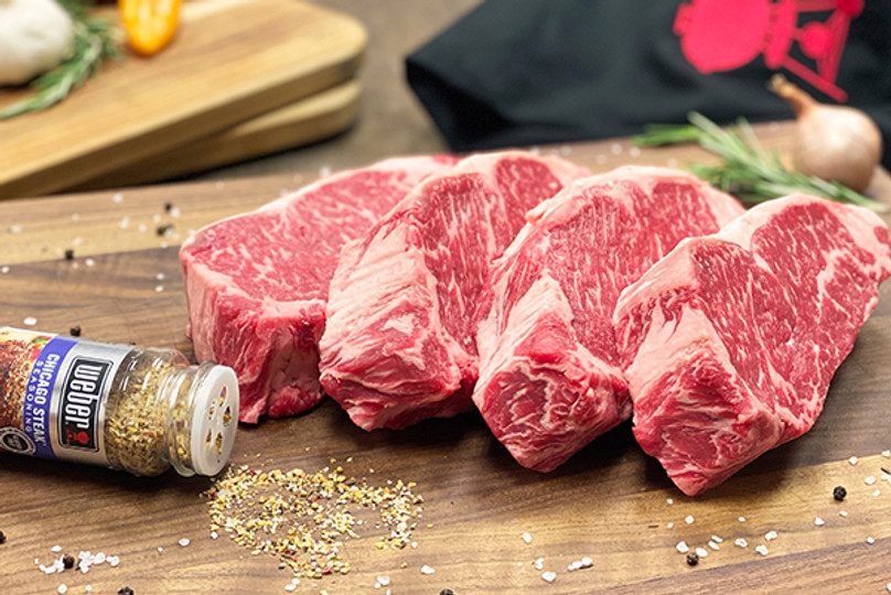 Pictured are 4 uncooked Linz Heritage Angus boneless ribeyes with a container of seasoning on a cutting board.