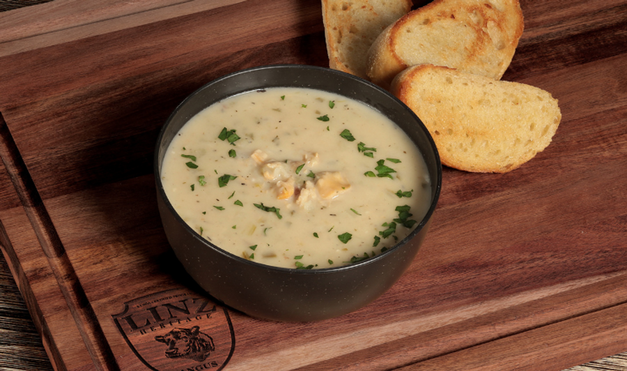Pictured is our LINZ Clam Chowder topped with herbs in a bowl on a table with clams, bread in the background