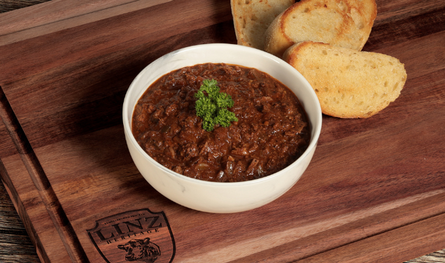 Pictured is our LINZ Spicy Texas Beef Chili topped with herb garnish on a wooden table with toasted bread in the background.