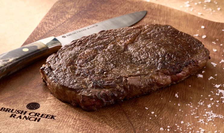 Pictured is a cooked Prime wagyu boneless ribeye from Brush Creek Ranch on a wooden cutting board with a Brush Creek branded knife