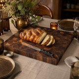 Large 24x24 walnut end grain cutting board styled on a dining table with sliced challah bread and bread knife