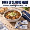 Crystal Hot Sauce 7 gram Packets 200 Ct, image of hot sauce packets on a wooden table alongside a bowl of shrimp gumbo garnished with rice and green onions, with bread on a side.