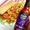 Bayou Gotham Ruby Rebelle Bourbon Cayenne Hot Sauce 5oz, a bottle of hot sauce with purple and black label featuring flame graphics, held in front of a shrimp po' boy sandwich with lettuce, tomato, fried shrimp, and French fries.