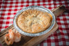 La Boucherie Shrimp Pie, a 0.75 lbs savory seafood pie in a disposable aluminum container on a wooden board, with a cooked shrimp next to it, placed on a red and white checkered tablecloth.