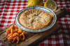La Boucherie Crawfish Pie, 0.75 lbs, in a foil container on a wooden cutting board, served with a side of seasoned crawfish and fresh lemon halves on a red checkered tablecloth.