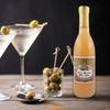 Boscoli Dirty Martini Olive Juice 12.7oz 12 Pack, a bottle of olive juice with a label, beside cocktail glasses with green olives garnished with toothpicks, and a small glass filled with green olives on a rustic wooden surface.