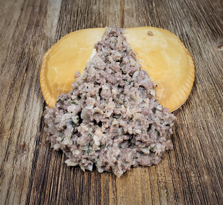 Mrs Wheat's Mild Meat Pies 48ct 4oz, image showing a meat pie with a golden crust and a pile of cooked ground meat filling on a rustic wooden surface.