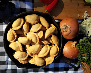 Mrs Wheat's Mini Spicy Meat Pies 16ct 1oz, a skillet filled with small, golden, seasoned meat pies on a checkered tablecloth.