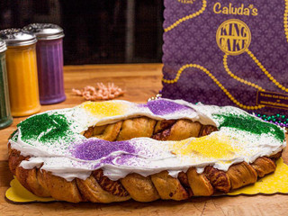 Creole Foods of Louisiana Caluda's Traditional King Cake, decorated with colorful icing and sprinkles, with a purple box in the background.