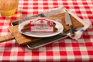 The Best Stop Smoked Boudin 12oz, packaged smoked boudin sausage on a white plate on a wooden cutting board with a knife, set on a red and white checkered tablecloth.