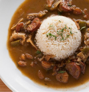Poche's Poches Chicken & Sausage Gumbo 2lb, bowl of hearty gumbo with chicken, sausage, and vegetables served with a mound of rice garnished with herbs.