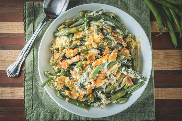 La Boucherie Green Bean Casserole 2 lb, served on a white plate with melted cheese and sliced almonds, garnished with spices, on a green napkin and wooden table.