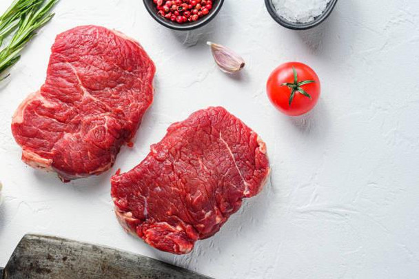 8oz Angus Beef Filet (Pack of 2), fresh raw beef filets on a white surface with cherry tomato, garlic, black pepper, and herbs in the background.