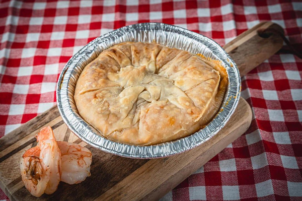 La Boucherie Shrimp Pie, a 0.75 lbs savory seafood pie in a disposable aluminum container on a wooden board, with a cooked shrimp next to it, placed on a red and white checkered tablecloth.