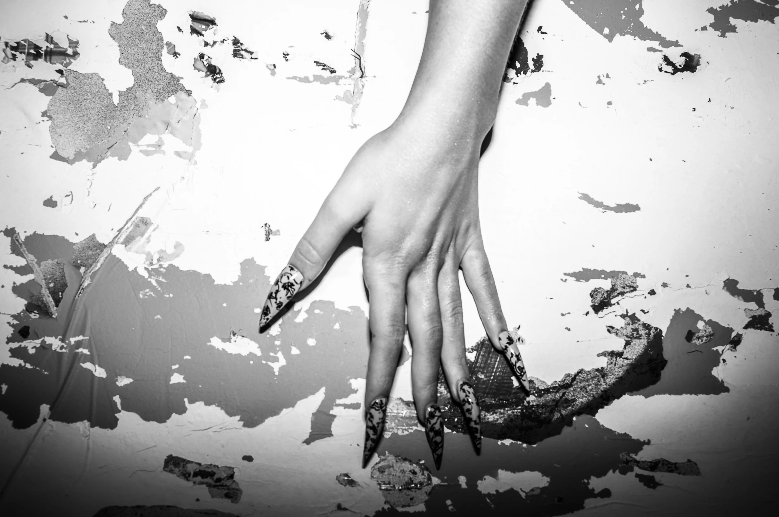 Black-and-white photograph of a hand with patterned nails against a textured wall