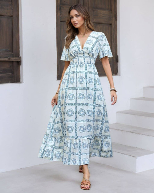 Model wearing a white and blue Mediterranean print maxi dress with sunburst patterns, V-neckline, flutter sleeves, and a smocked waist standing on white stairs.