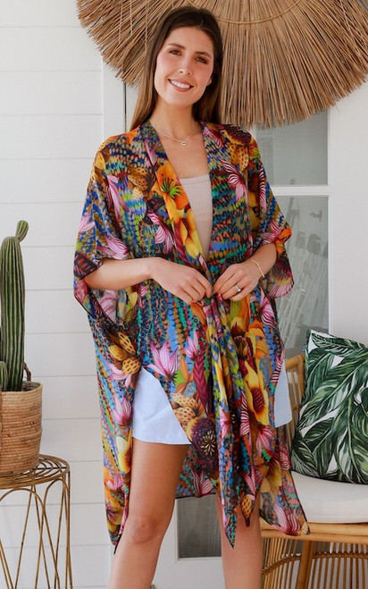 Model wearing the easy open-front Exotic Kaleidoscope Kimono. The colourful kimono shows a bright print of pink lilies and yellow sunflowers over a casual white top and denim shorts.